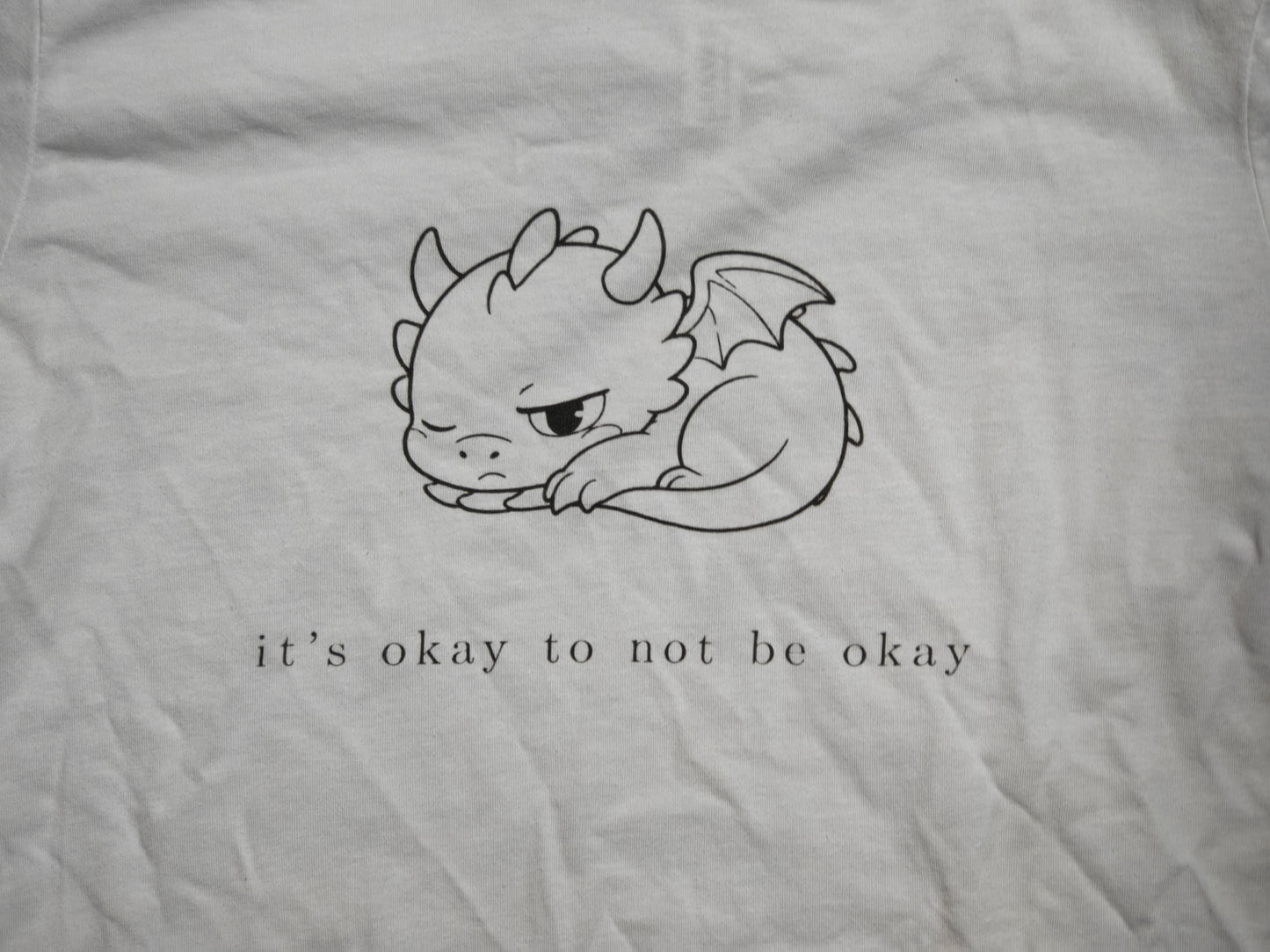 Macro close-up of the It's Okay To Not Be Okay line-art print on real white cotton, showing fine linework and visible fabric weave.